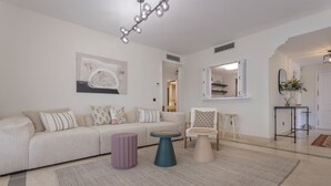 A modern living room with a sofa, coffee table, and chair. There is a large painting on the wall and a mirror above the door. - 3 Bedroom, 3 Bathroom Beachside Apartment (Estepona)