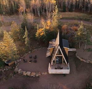 A cabin with a triangular roof, surrounded by trees and a gravel area with outdoor seating. - Cliffside A-Frame (Two Harbors)