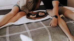 A couple sitting on a bed with a tray of food, in a room with a plain wall and a small decorative item. - Cliffside A-Frame (Two Harbors)