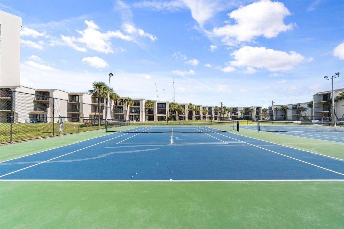 Ein Tennisplatz im Freien, umgeben von mehrstöckigen Gebäuden mit Balkonen, unter blauem Himmel.