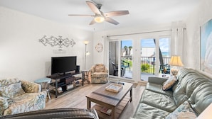A living room with a flat-screen TV, a ceiling fan, a sofa, a coffee table, and a balcony with outdoor furniture. - 3BR Sleeps 8 | Sugar Beach | Steps to Beach & Pool (Panama City Beach)