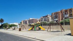 Ein Spielplatz mit Schaukeln und einer Rutsche, umgeben von einer niedrigen Mauer und Gebäuden im Hintergrund.