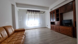 Ein Wohnzimmer mit braunem Ledersofa, einer Holzkommode mit Fernseher und einem Fenster mit transparenten Vorhängen.