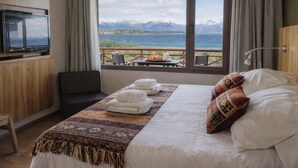Habitació d'hotel amb llit, televisió, vista a muntanyes i un llac, i taula de petit esmorzar amb menjar.