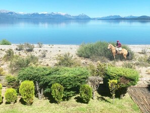 Una persona monta a caballo por un sendero junto al lago, con montañas al fondo.