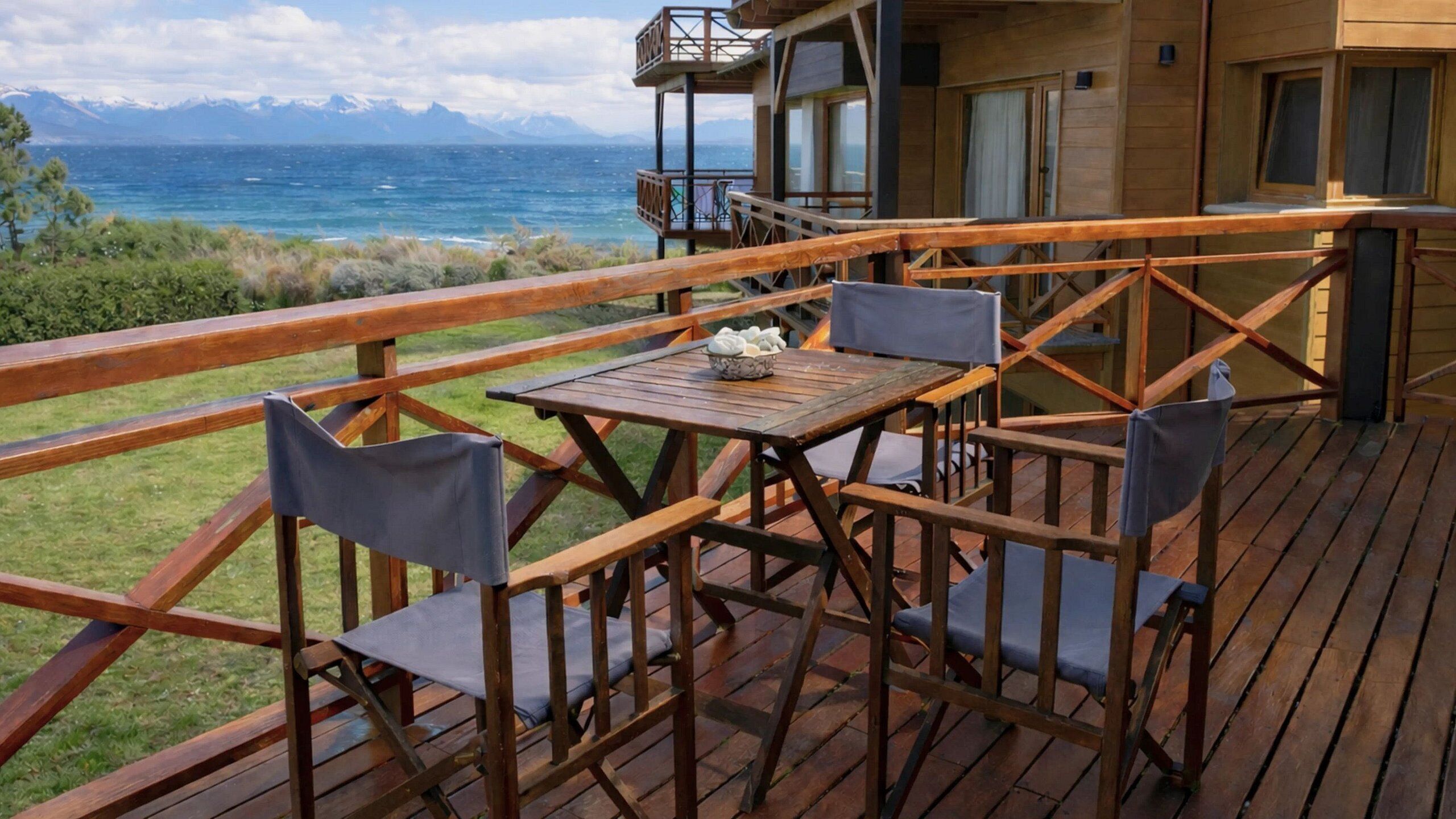 An outdoor deck with wooden furniture and a table, overlooking a body of water and mountains.