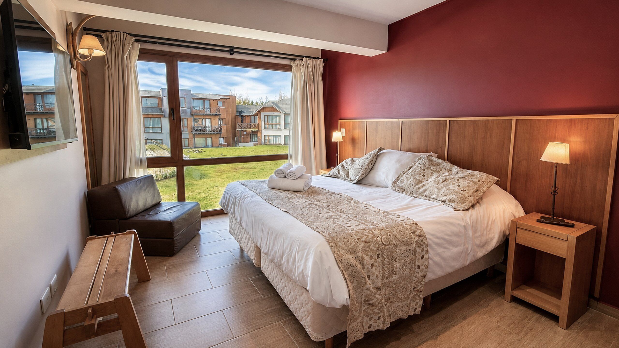 A hotel room with a large bed, a wooden headboard, a bedside table, a desk, and a view of a building and greenery outside.
