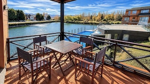 Een houten terras met een tafel en stoelen, met uitzicht op een rivier, met een zwembad en gebouwen op de achtergrond.