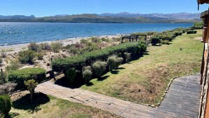 Una terraza de madera que da a una playa, con vista a un lago y montañas a lo lejos.