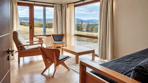 A room with a bed, two chairs, a small table, and a view of a mountainous landscape through large windows. - Garden Superior Moreno Ground Floor Lake Front (Dina Huapi)