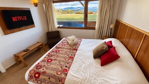 Habitación de hotel con cama, banca, televisor montado en la pared y ventana con vista a montañas.