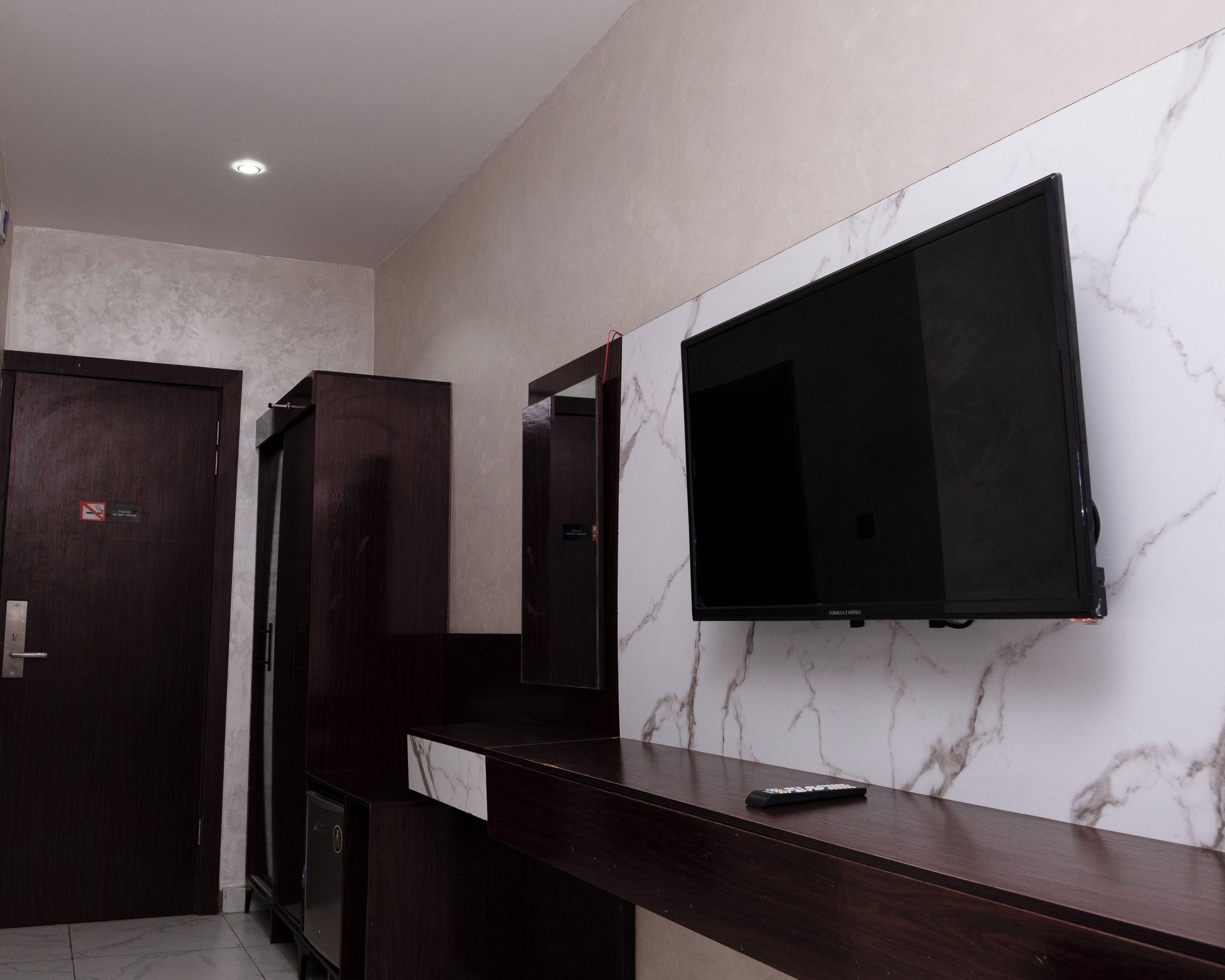 A modern hotel room with a large flat-screen TV mounted on a marble wall, a wooden counter, and a door with a no-smoking sign.