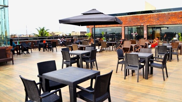 An outdoor patio with tables and chairs, a large umbrella, and a brick building in the background.