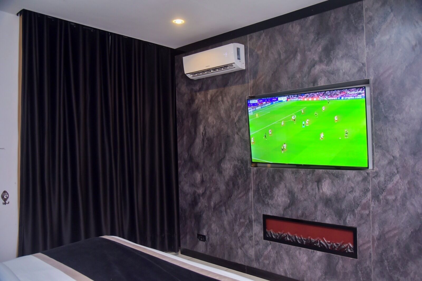 A room with a mounted TV displaying a football match, a wall-mounted air conditioner, and a bed with a dark headboard.