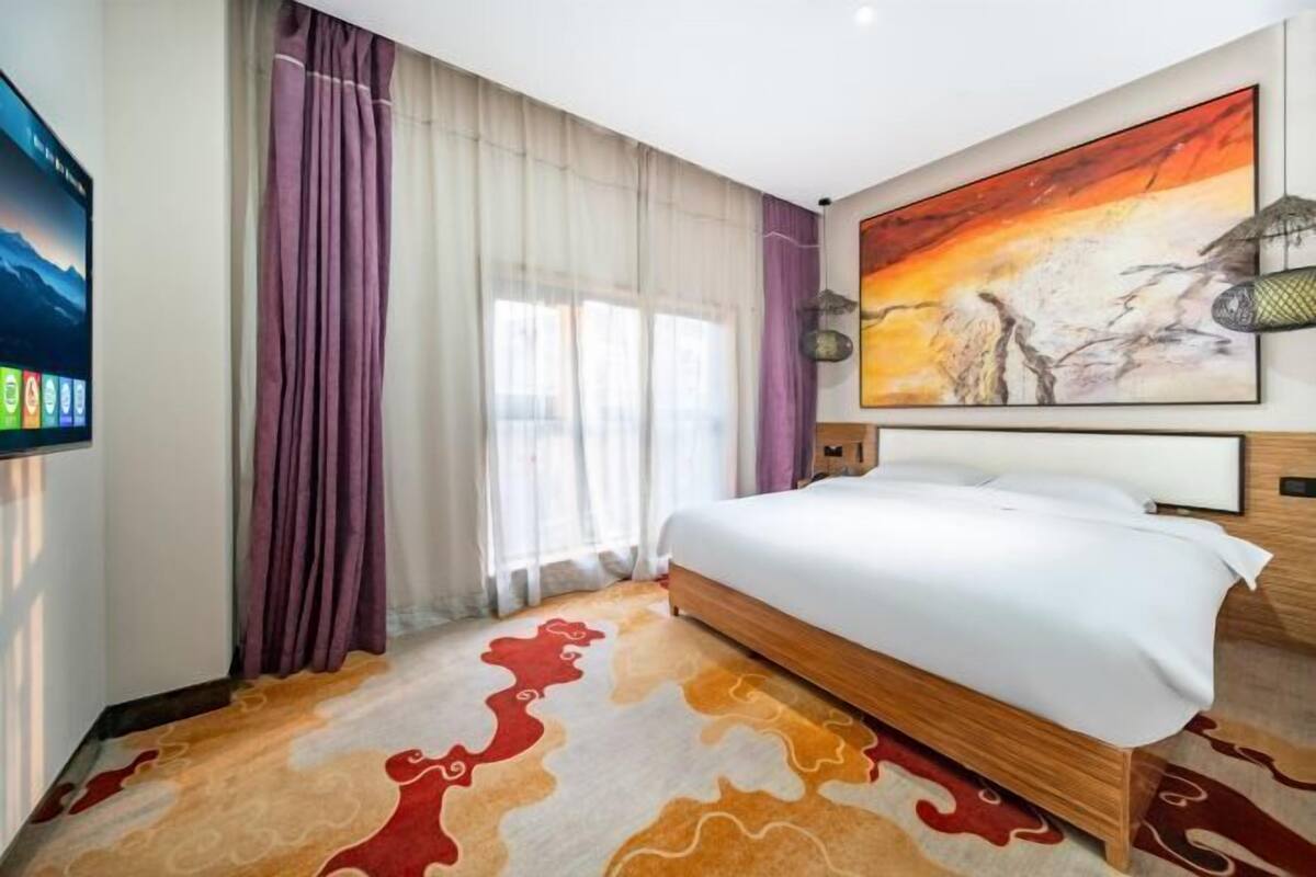Habitación de hotel con cama, cortinas, un cuadro, televisor y alfombra estampada.