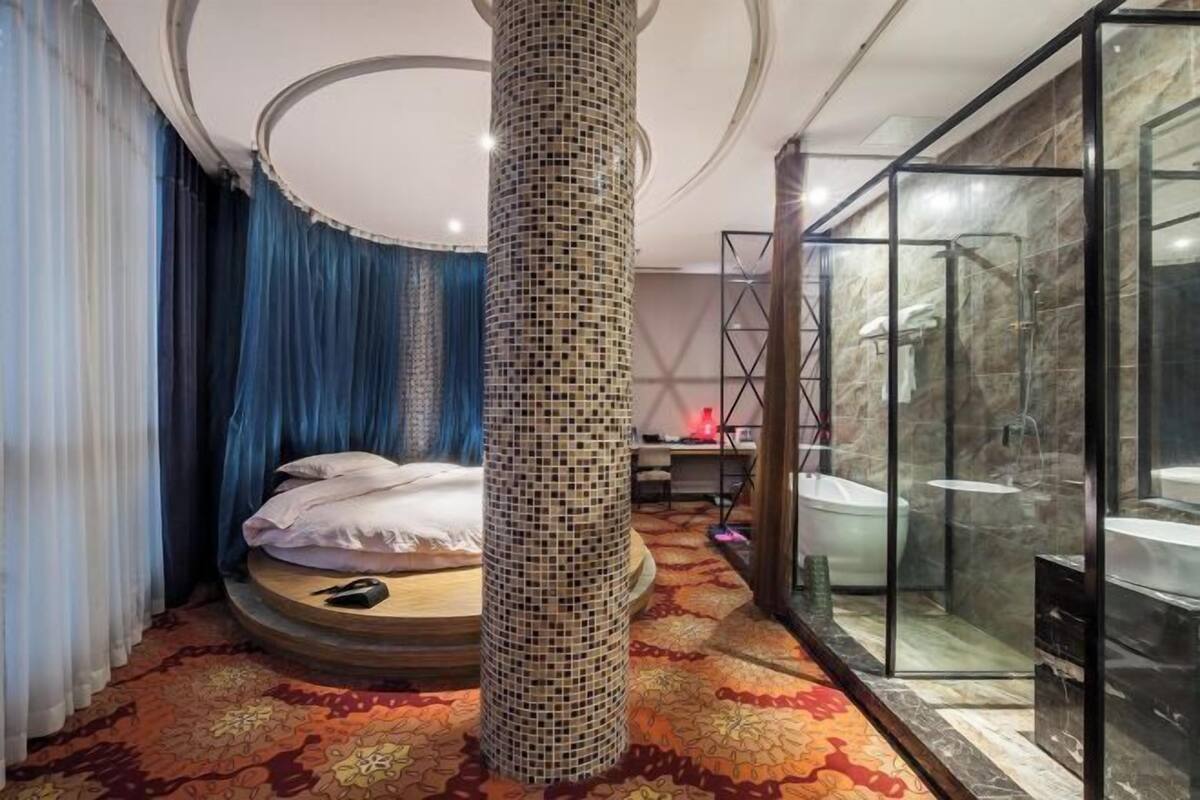 Una habitación de hotel con una cama redonda, un pilar decorativo, una ducha con paredes de vidrio y una alfombra estampada.