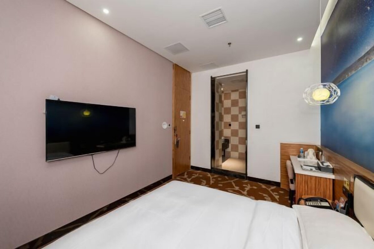 Habitación de hotel con cama, televisor montado en la pared, escritorio con silla y baño visible a través de una puerta abierta.