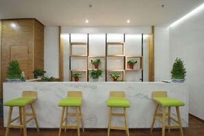 A reception area with a marble counter, wooden chairs with green cushions, and potted plants. - Aifeier Shishang Hotel (Zhengzhou)