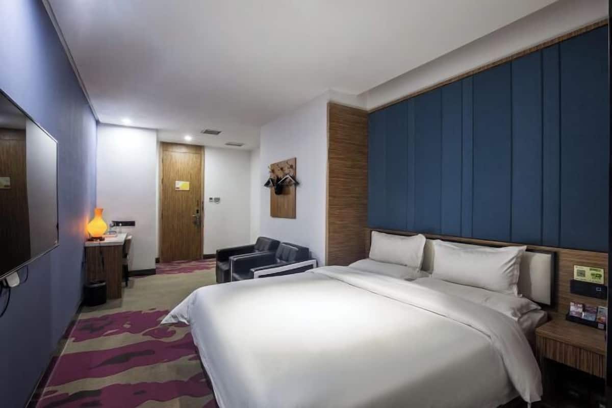 Habitación de hotel con una cama grande, un escritorio con lámpara, una silla y una puerta de madera.