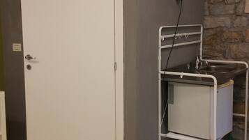 Una habitación con una puerta blanca, una kitchenette pequeña con pileta y espacio de almacenamiento, una pared de piedra y un tapete estampado.