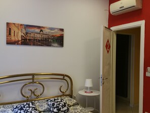 A bedroom with a bed, a wall-mounted air conditioner, a door with a number, and a small table with a lamp. - Guerrero Rooms (Trieste)