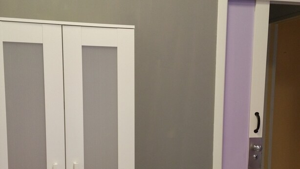 Une pièce avec une armoire blanche, un lit recouvert d’une serviette rose et violette, et une porte ornée d’un panneau violet.