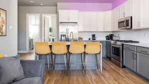 A modern kitchen with white cabinets, a stainless steel refrigerator, a microwave, and a gas stove. There are four bar stools at the island. - Tailgater's Dream! Rooftop w/ TV + Fire + Lights (Nashville)