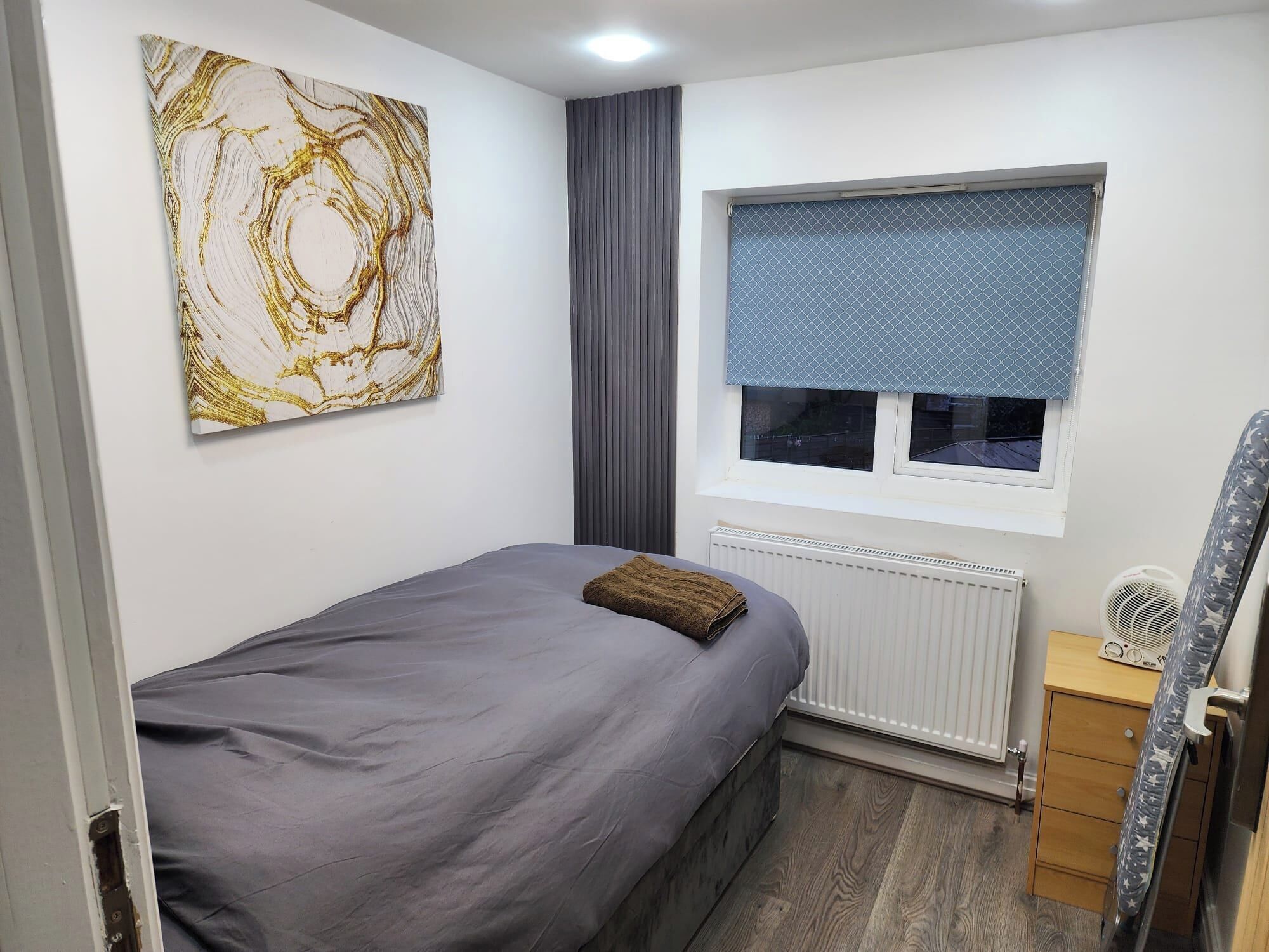 A bedroom with a bed, a radiator, a window with blinds, a radiator cover, a wooden dresser, and a star-patterned chair.