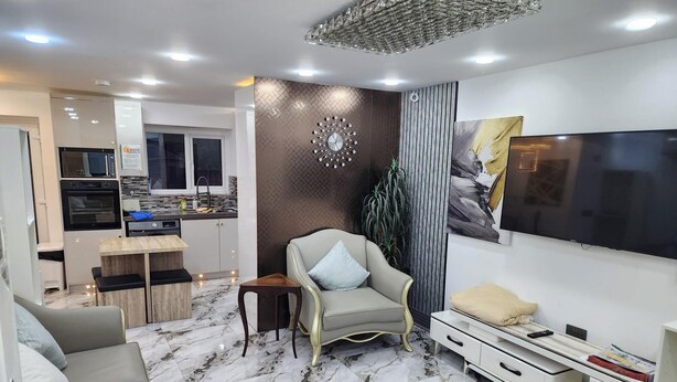 Un salon moderne avec un canapé, un téléviseur, un élément décoratif mural et, au fond, un coin cuisine.