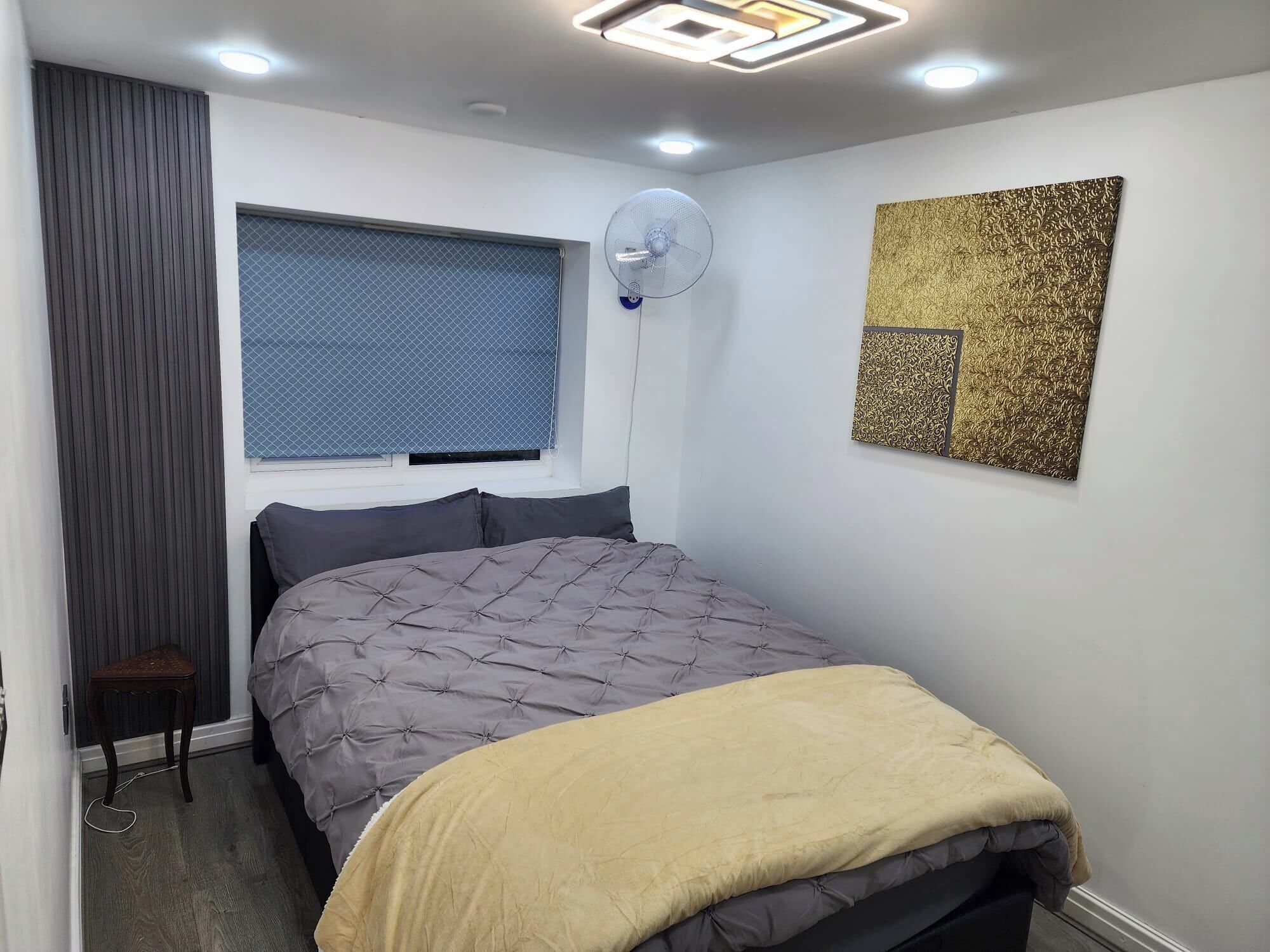 Un dormitorio con cama, ventilador de pie, ventana con persianas y un adorno en la pared.
