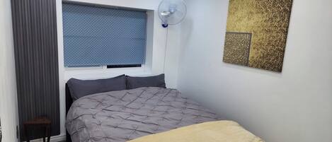 A bedroom with a bed, a standing fan, a window with blinds, and a decorative wall piece.