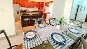 Un coin repas avec une table dressée pour quatre personnes, une cuisine en arrière-plan et une plante en pot sur la table.