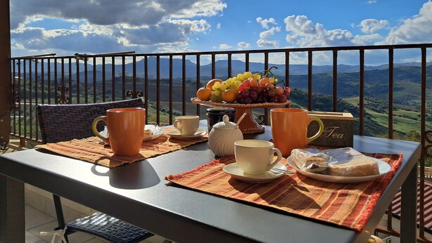 A balcony with a table set for breakfast, including a fruit platter, cups, and a bread roll.