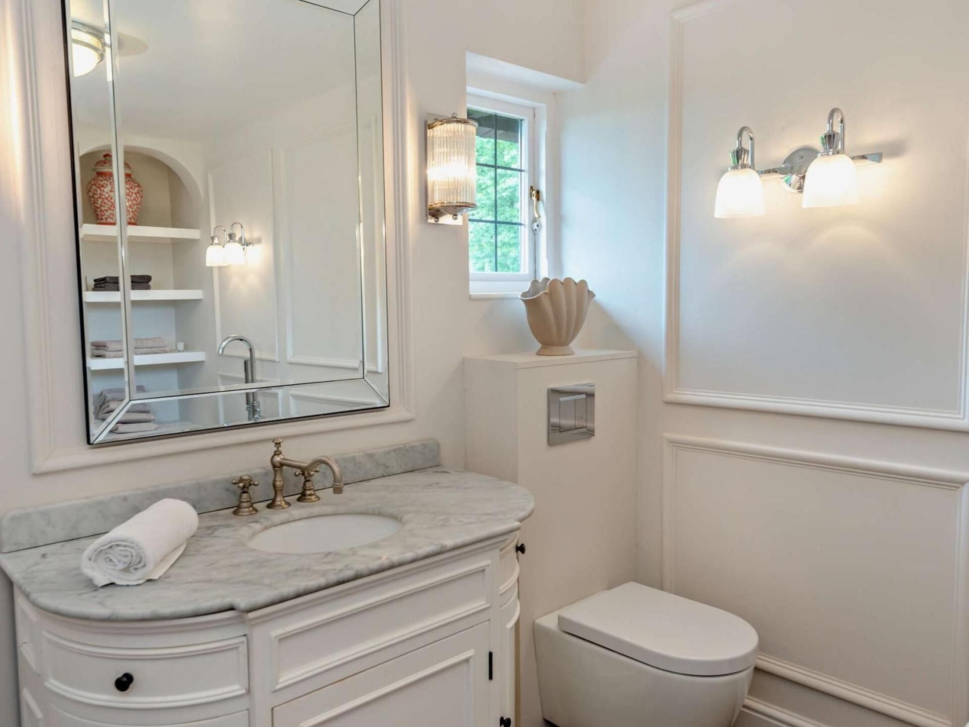 A bathroom with a white vanity, a sink, a mirror, a toilet, and wall-mounted lighting.