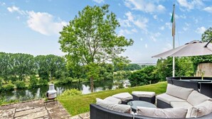 Une terrasse dotée de meubles en osier, d’un chemin de pierre et offrant une vue sur un lac et des arbres.