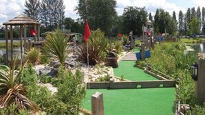 Een midgetgolfbaan met een groen oefenveld, houten constructies en verschillende planten.