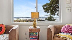 Una habitación con un ventanal grande que ofrece vistas al mar y árboles, una mesita de madera con libros, y dos sillones con cojines.
