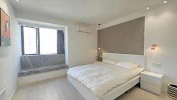 Une chambre avec un grand lit, un banc près de la fenêtre, une table de chevet et un luminaire fixé au mur.