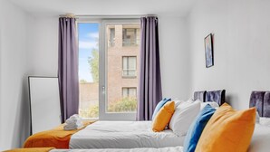 A hotel room with two beds, a window with curtains, a flat-screen TV, and a view of a brick building. - Modern 2-bed Apartment in Stratford (London)