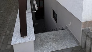 A narrow outdoor corridor with a metal railing, a small window with blinds, and a door leading to a darker interior.