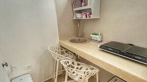 Una pequeña kitchenette con un lavamanos, dos sillas blancas, una encimera y una ventana arriba.