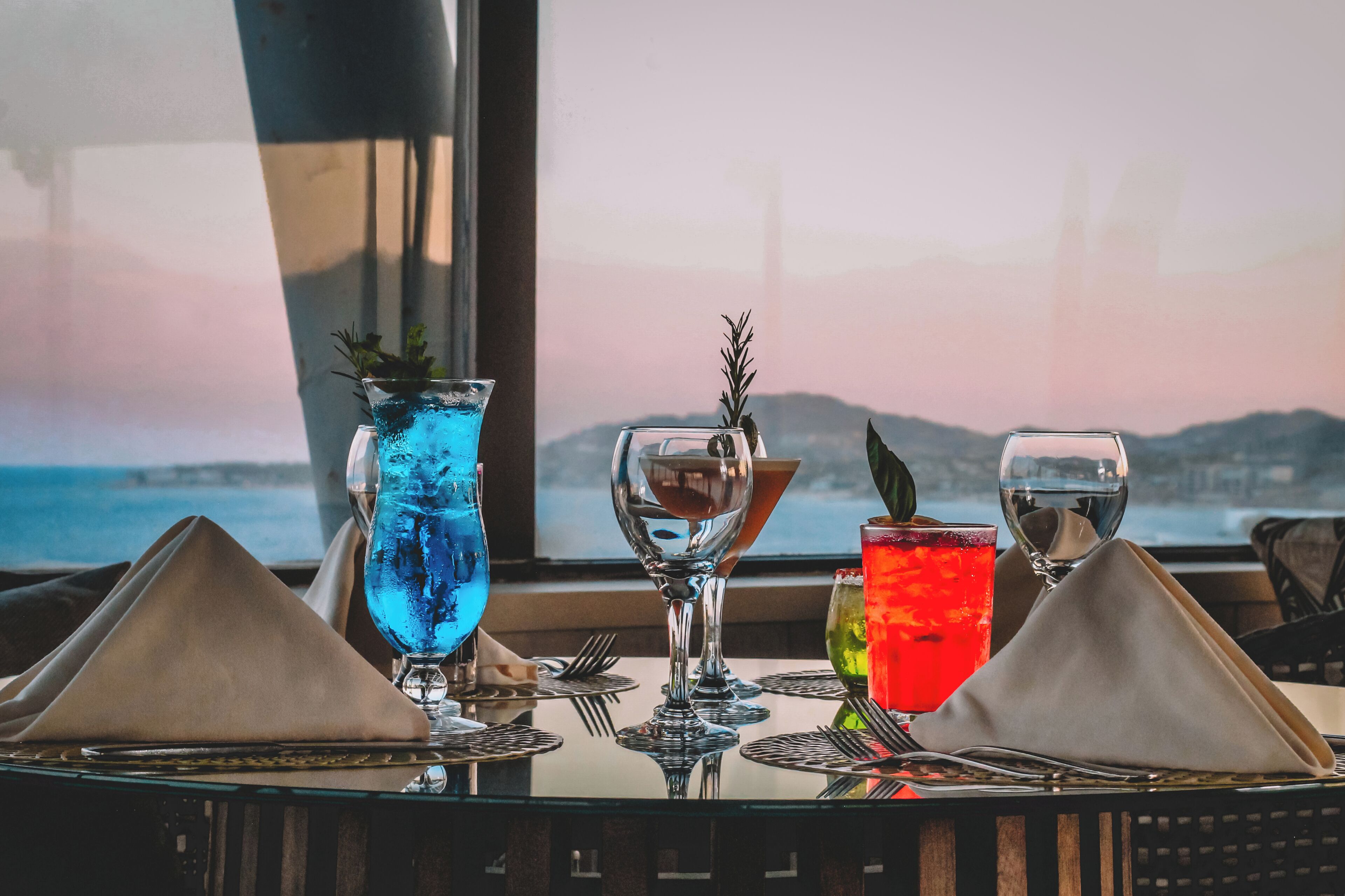 Una mesa con tres vasos de bebidas, una copa de vino y vista al mar a través de una ventana.