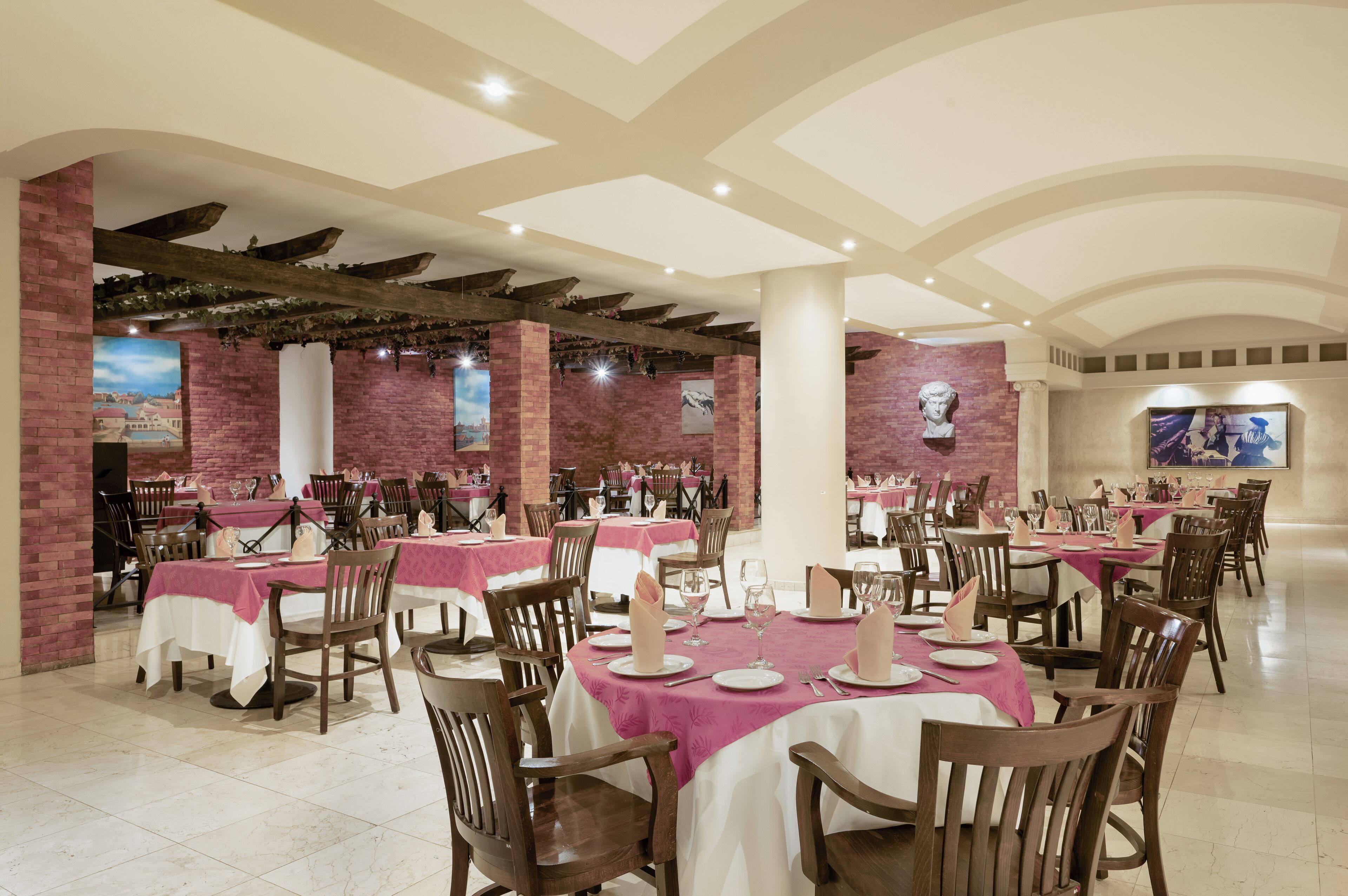 A restaurant with round tables covered in white tablecloths and pink napkins, wooden chairs, and brick walls.