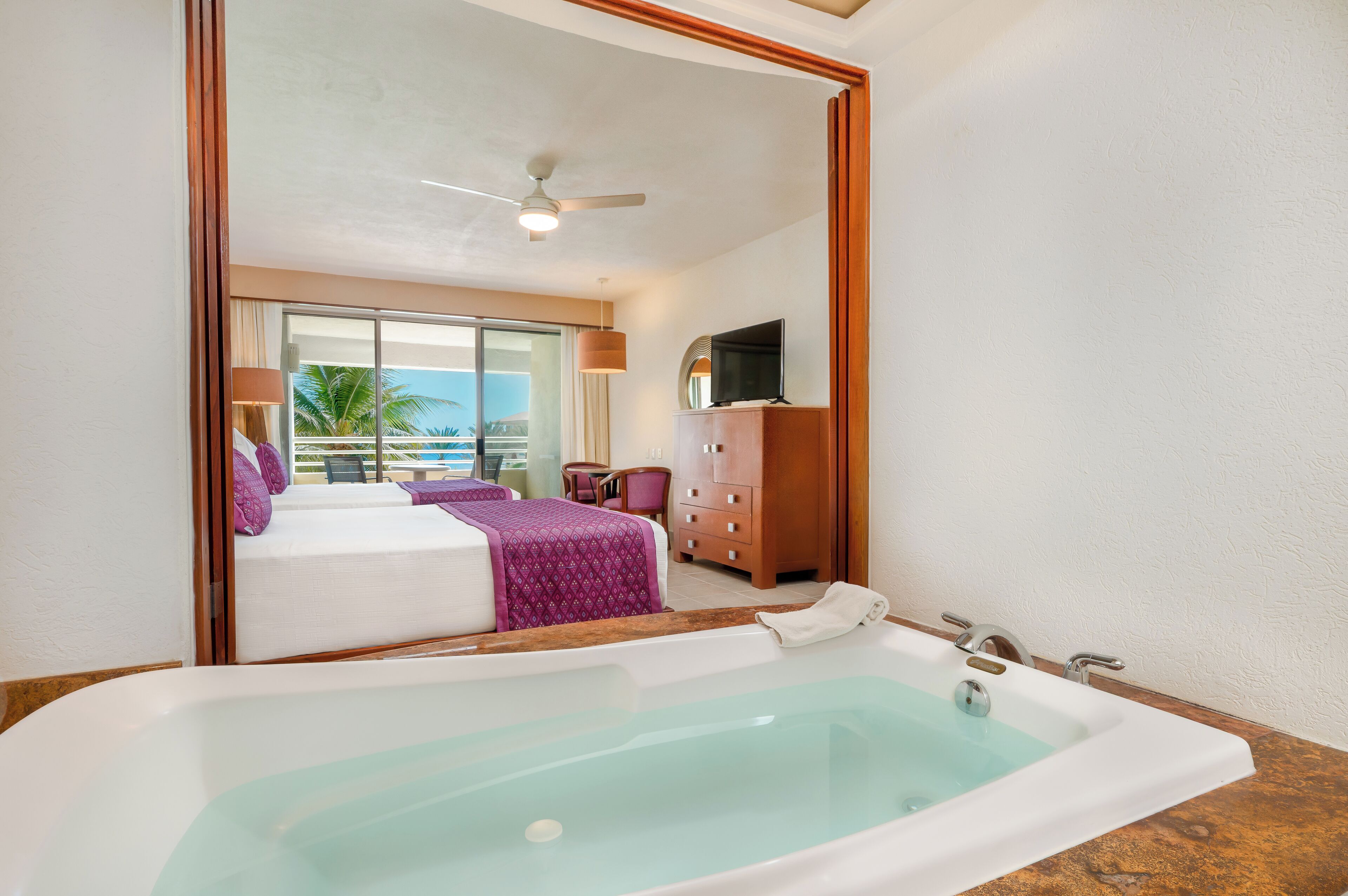 A hotel room with a large bathtub, a bed with purple bedding, a TV on a wooden cabinet, and a view of the ocean through a sliding door.