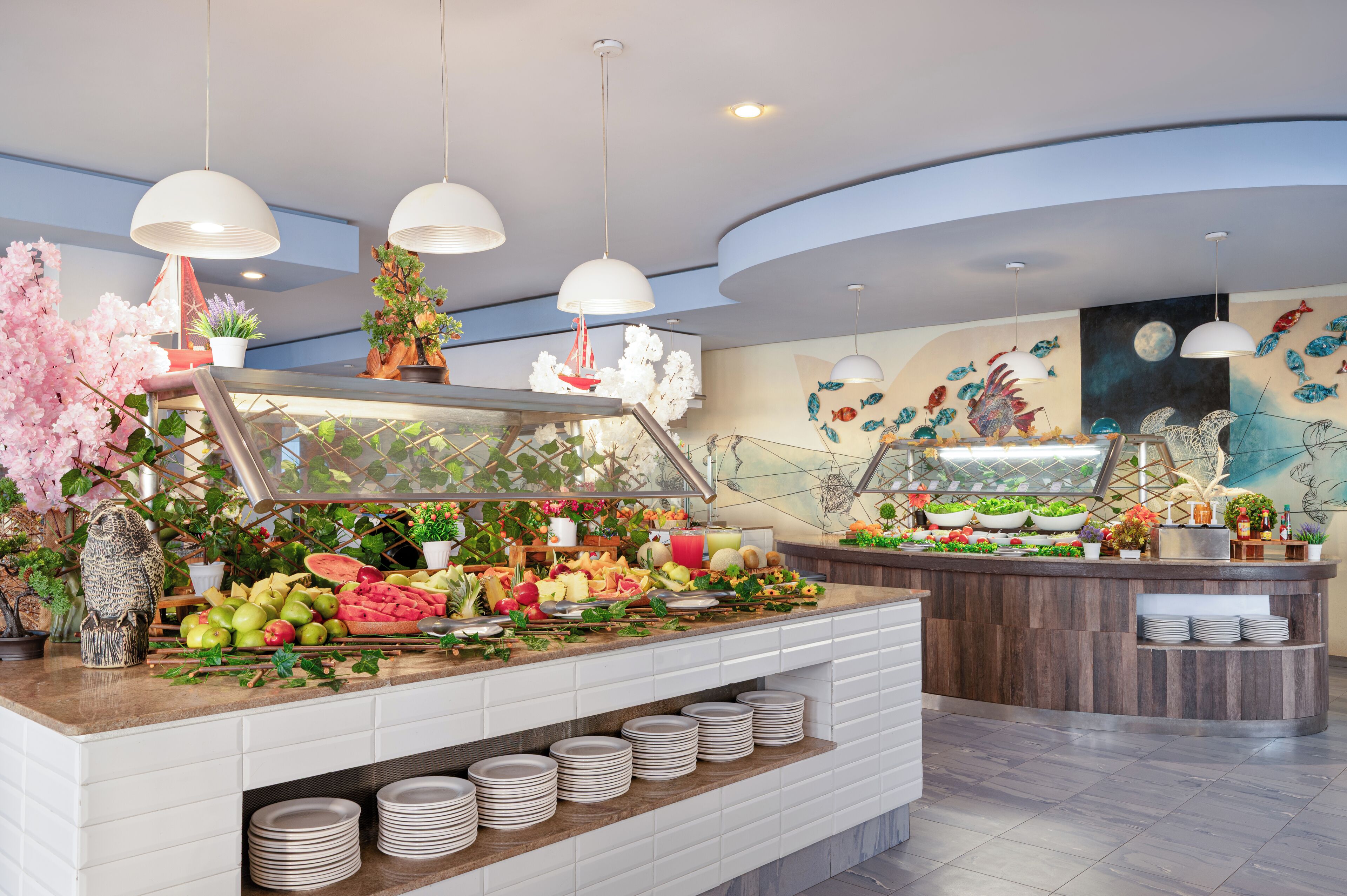 A buffet area with a variety of fruits and vegetables, a selection of beverages, and a decorative wall with fish and moon mural.