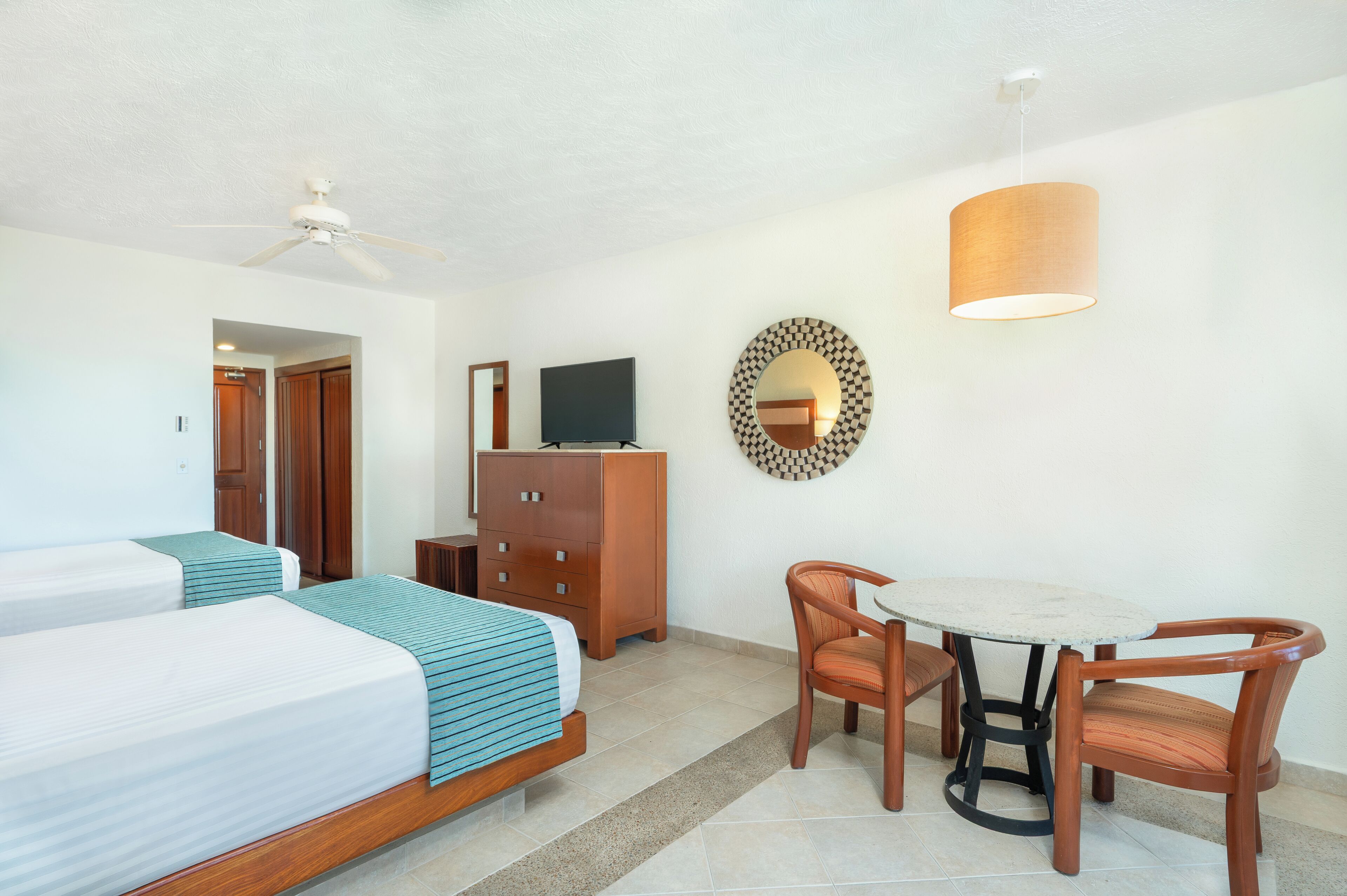 Een hotelkamer met twee bedden, een televisie, een dressoir, een spiegel, een ronde tafel met stoelen en een plafondventilator.