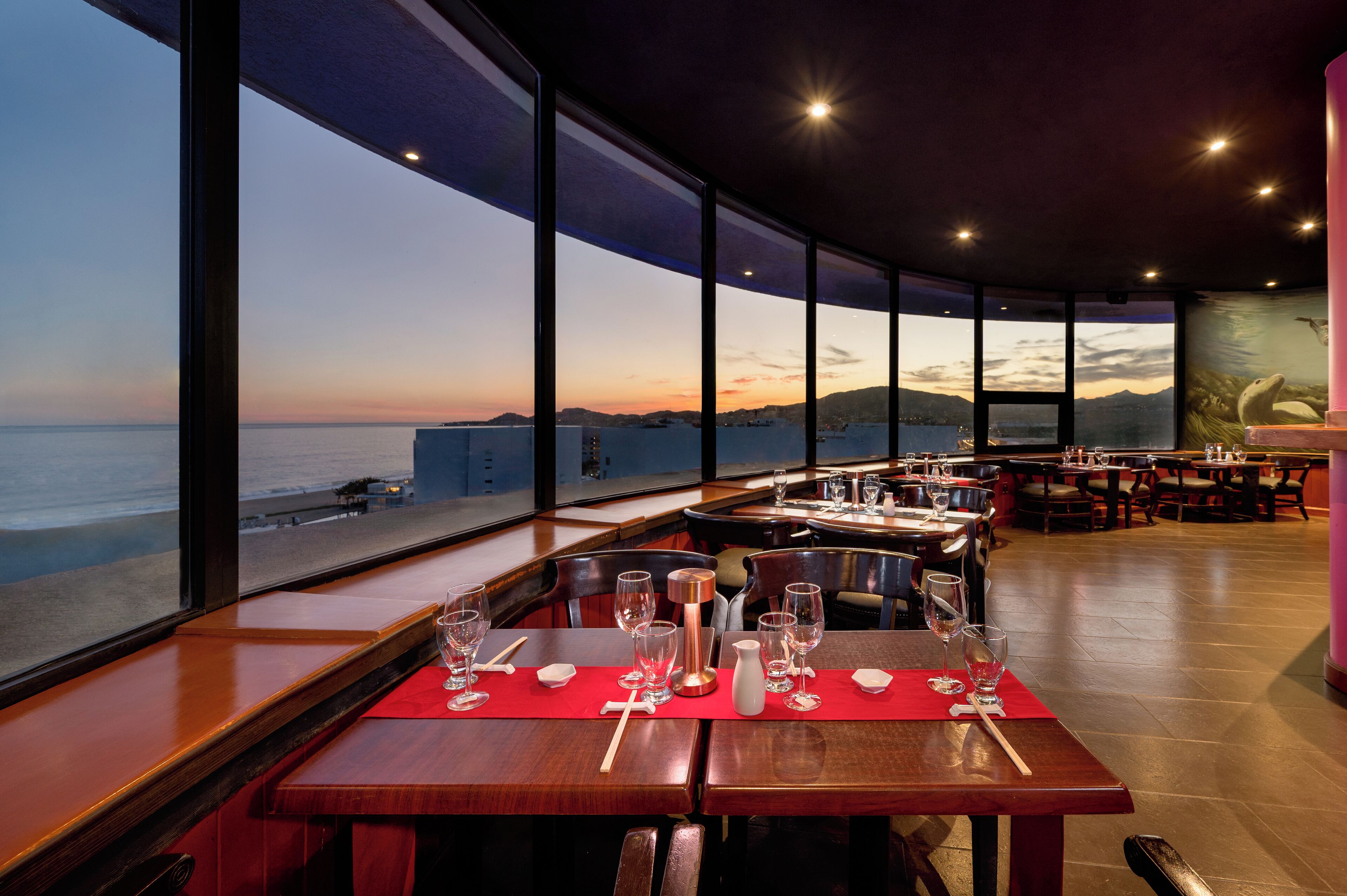 A restaurant with a view of the ocean at sunset, featuring wooden tables and red placemats.