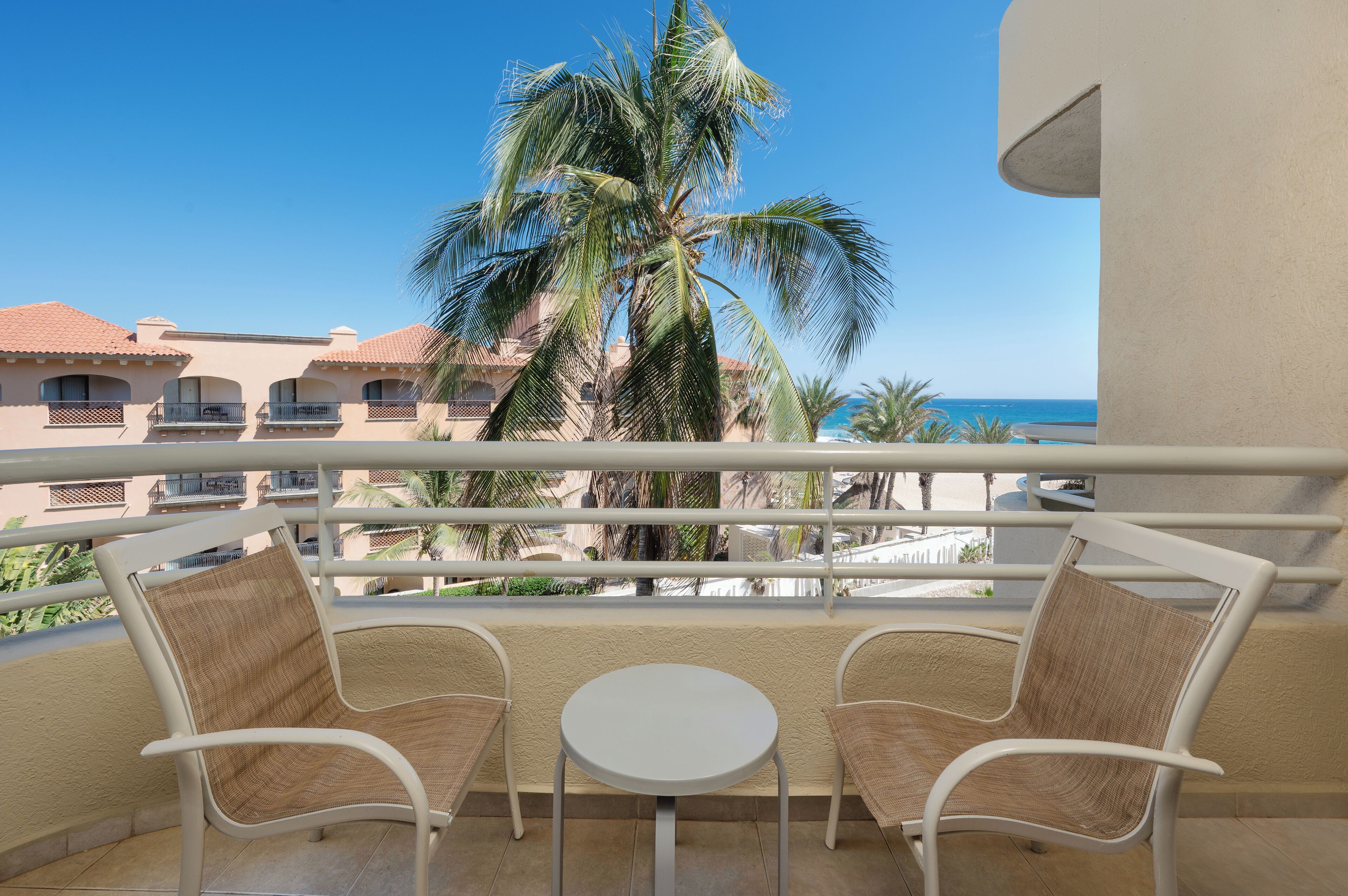 Un balcon avec deux fauteuils en osier et une petite table ronde, d’où l’on peut admirer un palmier et une plage.