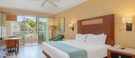 A hotel room with a large bed, a desk with a lamp, a chair, and a view of palm trees through the window.