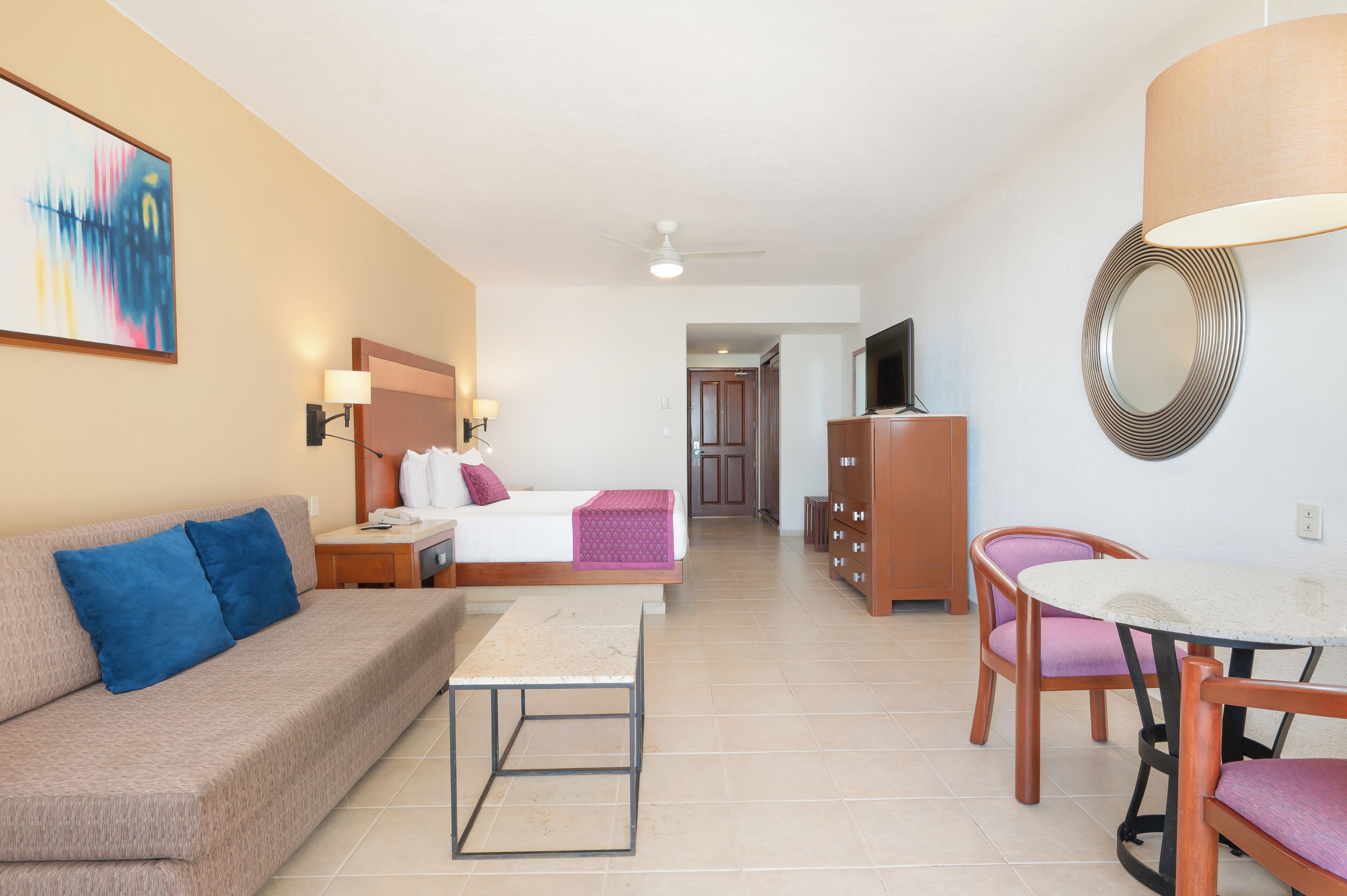 A hotel room with a sofa, a bed, a small table, a dining table with chairs, a TV, a mirror, and a painting on the wall.